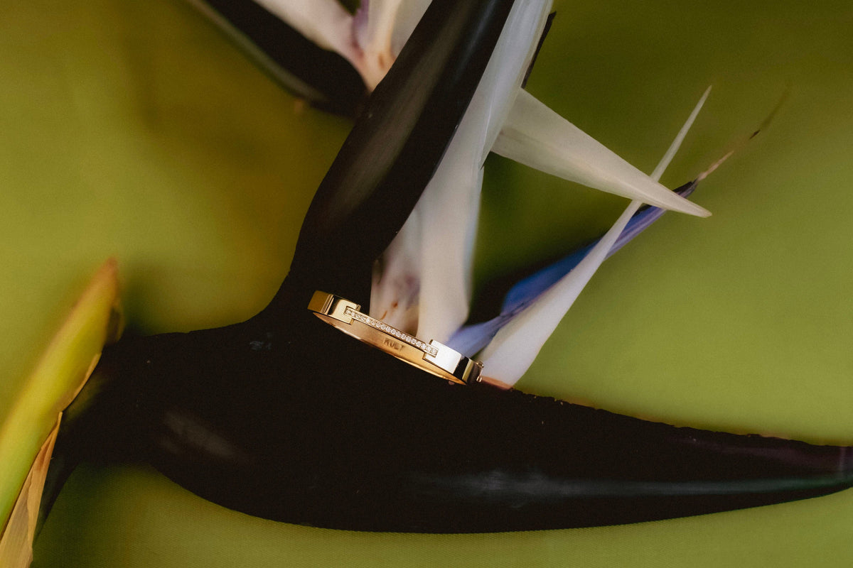 KULT La Ligne bangle bracelet ring with diamond-set bar resting on a dark bird-of-paradise flower against an olive green background