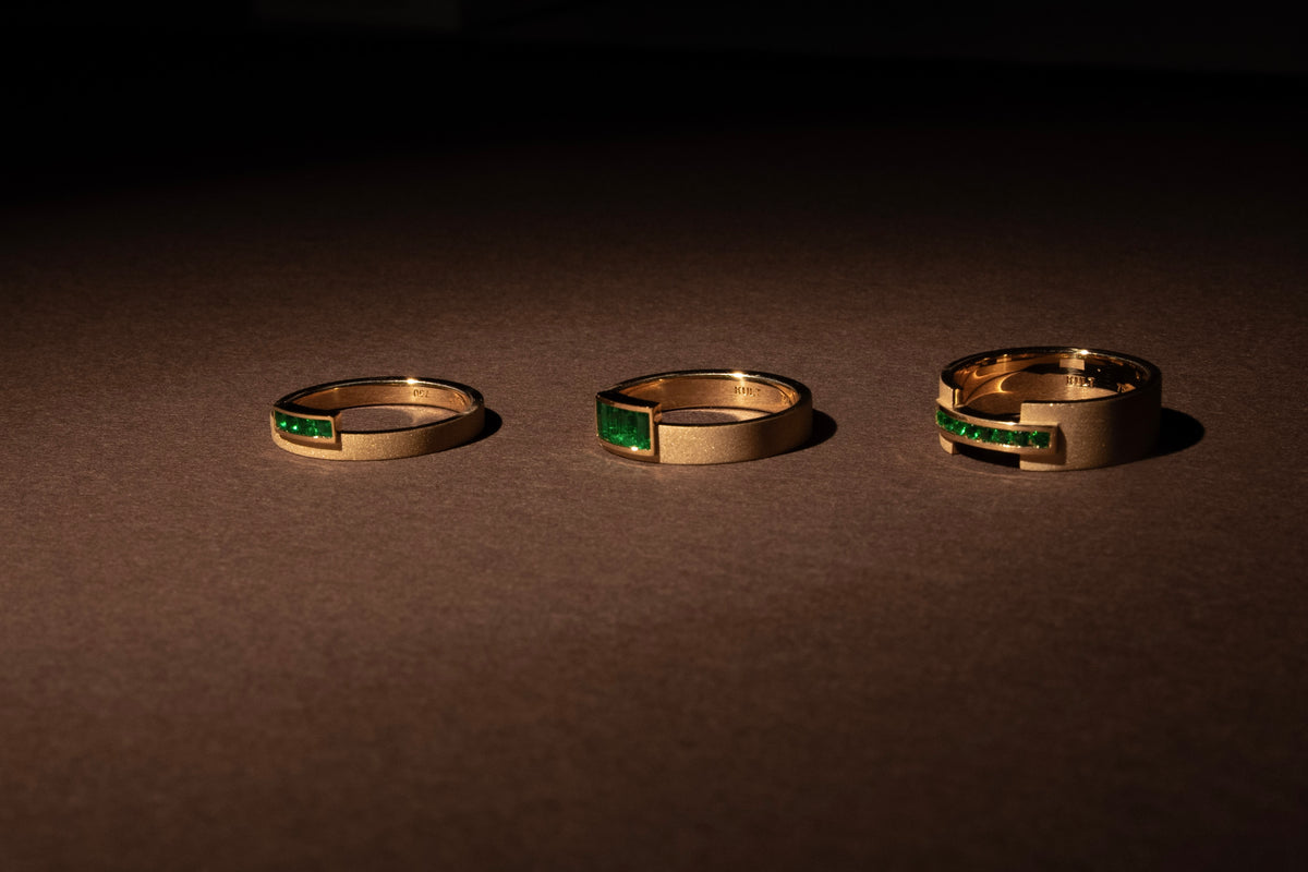 Three custom gold rings with emerald accents arranged on a dark surface in dramatic light