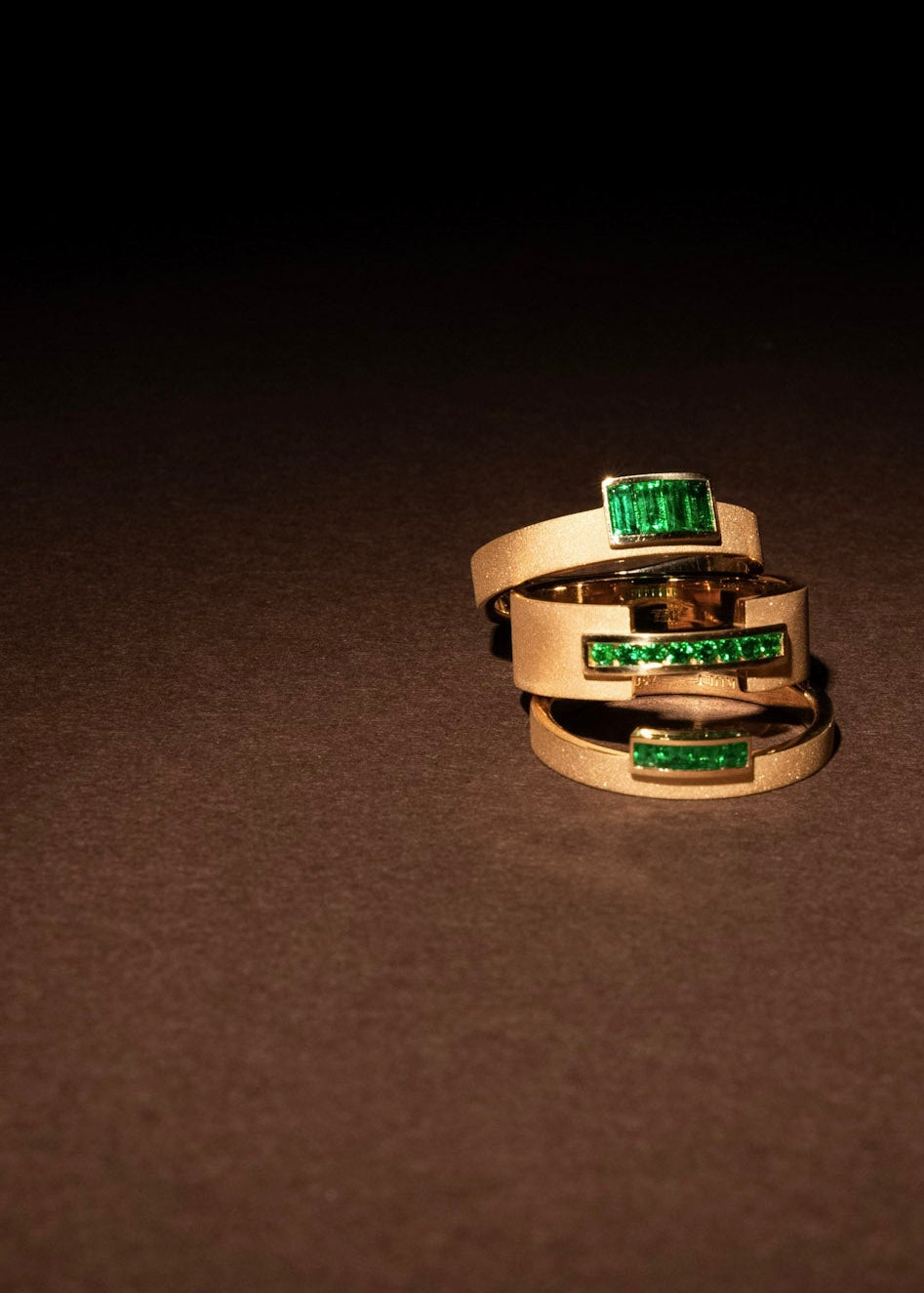Three gold rings with emerald accents arranged on a dark surface in dramatic light