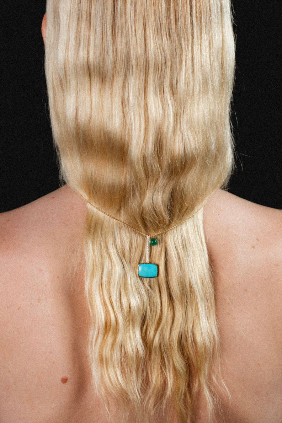 Gold chain necklace with a turquoise diamonds and emerald pendant, worn down the back of a woman with long blonde hair