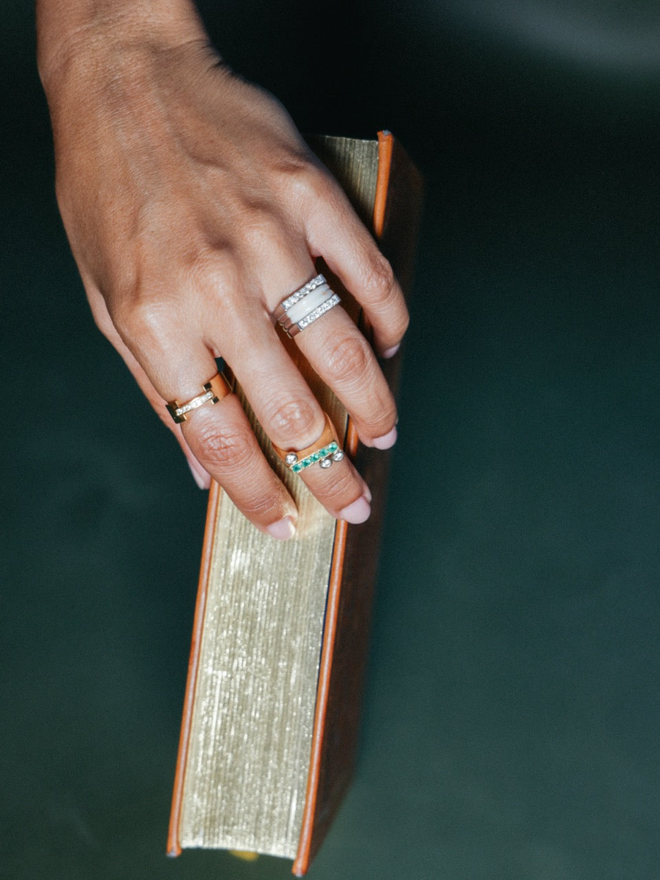 Hand wearing three gold rings with diamonds and emeralds, resting on a vintage book