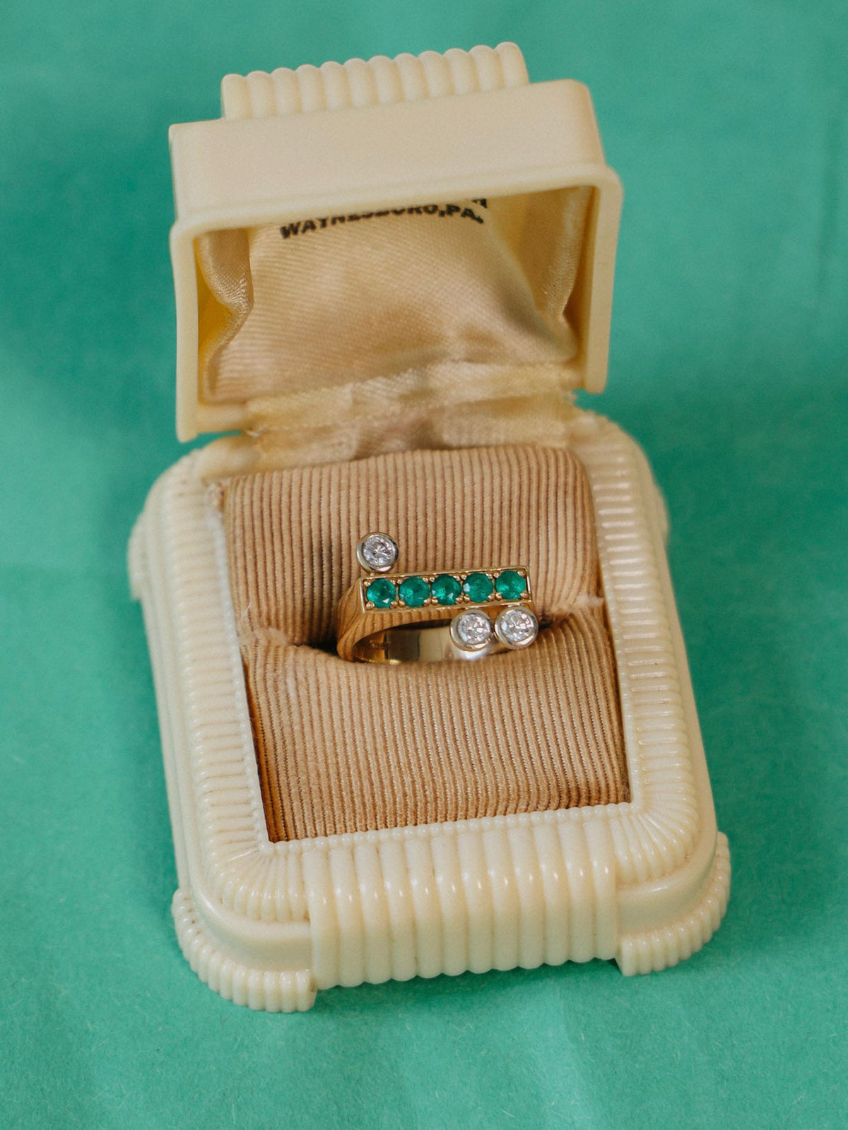 Clé N.1 yellow and white gold ring with emeralds and diamonds, placed  in an open vintage cream ring box standing on a green paper